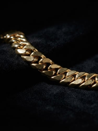 Wide panoramic shot of a heavy gold link bracelet on a matte black velvet surface, dramatic low-key studio lighting, sharp metallic gold highlights glinting from the left third, emerging from deep shadows, expansive dark background.