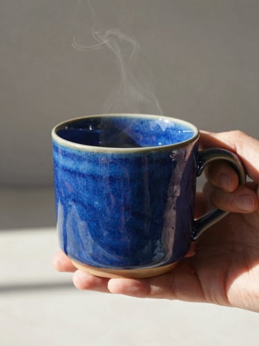 Warm editorial photography, a hand holding a steaming cobalt blue ceramic mug, bright natural sunlight casting soft shadows, glossy glaze texture, shallow depth of field, 35mm