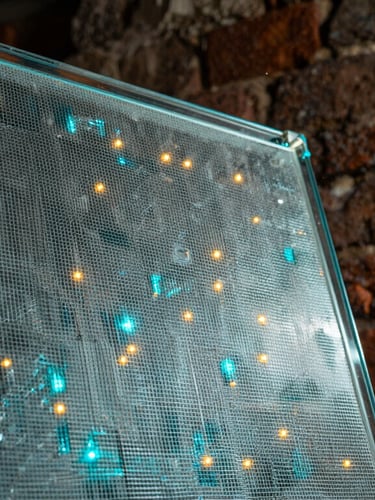 Macro detail of transparent micro-mesh LED film applied to a glass pane, glowing cyan and amber diodes active, historic Roman brick wall blurred in the background, dramatic side lighting.