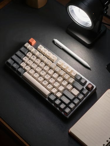 Overhead flat-lay on a dark desk surface: a compact mechanical keyboard, a precision stylus, and a closed notebook arranged in a tight cluster, lit by a single overhead studio strobe that reveals texture and material quality — no hands, no props, no distractions