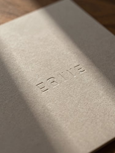 Close-up macro shot of embossed stationery on heavy cotton paper, soft indirect window daylight casting gentle shadows, warm walnut tones.