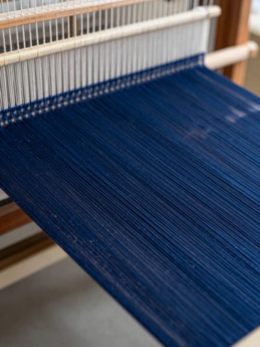 Macro close-up of deep indigo threads on an industrial loom, natural workshop light highlighting the fine texture of the warp and weft, professional textile manufacturing setting.