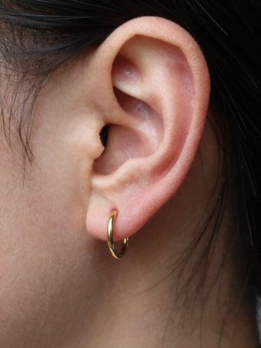 Close-up of a helix ear piercing with a small gold hoop, natural daylight from the side, skin detail sharp and clear, high contrast, clinical framing, no heavy editing