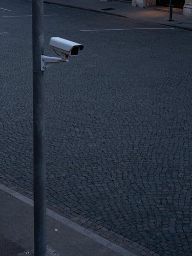 Corner-angle wide shot from above a dimly lit urban street intersection at dusk, a compact surveillance camera mounted on a concrete pole occupying the left third of the frame, empty cobblestone plaza stretching into the background, cool shadowless ambient light, no people, precise and calm framing mimicking a surveillance sightline