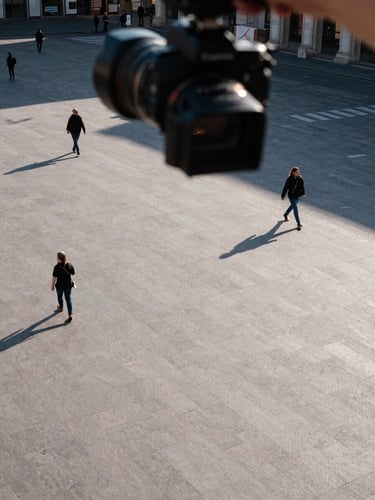 Corner-angle overhead shot looking down from a camera mount at a wide European public square, late afternoon daylight casting long precise shadows across paving stones, a few pedestrians mid-stride visible from above, the camera housing visible in the upper foreground as the compositional anchor, cool natural light, no faces identifiable