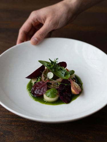 Moody culinary photography of hands plating a fresh artisanal dish, soft warm natural side-light, deep carmine and forest green tones, shallow depth of field, 35mm