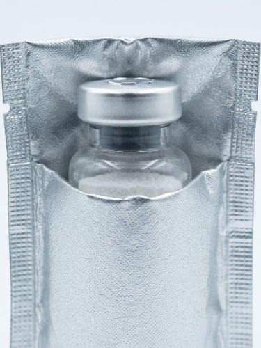 Crisp clinical product photography of a lyophilized peptide vial resting inside a custom sterile thermal foil insulation sleeve, cool silver tones, bright high-key studio lighting, white sterile background, macro lens, 50mm