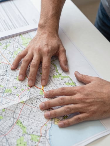 Hands spread over an OS map on a table, a finger tracing a field boundary, planning documents visible beside the map, clean LED indoor light, no faces