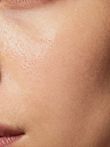 Extreme close-up of real skin texture showing pores and a natural luminous finish, soft gold highlighter catching indirect warm studio light