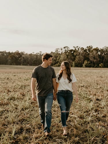Candid environmental portrait of a couple walking through an open field in North Carolina, soft overcast daylight, natural unposed interaction, muted earth tones