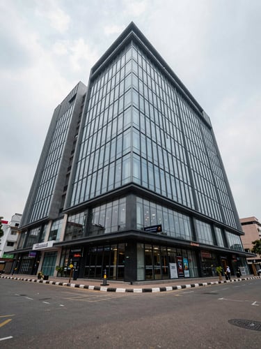 Wide exterior shot of a modern multi-storey commercial building in Vijayawada, photographed from street level looking up at the facade — overcast north-facing daylight, clean glass curtain wall, ground-floor retail signage visible, pedestrian footpath in foreground establishing urban zone context, no people posing
