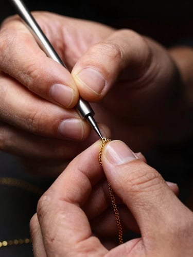 Close-up of a master jeweller's hands using a polishing tool on a delicate gold chain, warm studio light catching the metallic luster, dark background.
