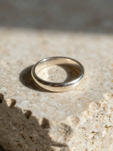 Macro close-up of a solid silver ring resting on a piece of raw, textured travertine stone, direct natural sunlight casting sharp architectural shadows