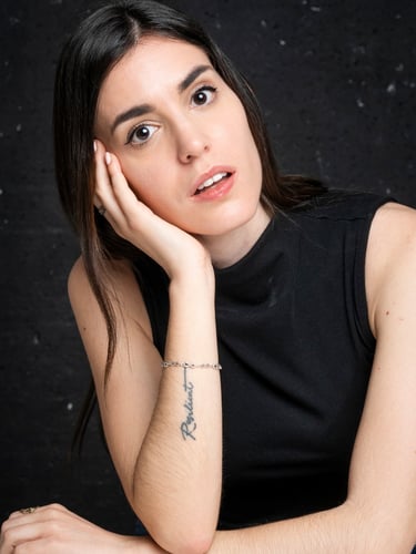 Professional headshot of a young woman with dark hair and a resilient arm tattoo posing against a dark background.