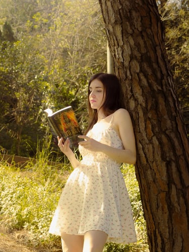A young woman in a floral summer dress leans against a pine tree reading a fantasy book in a sunlit forest.