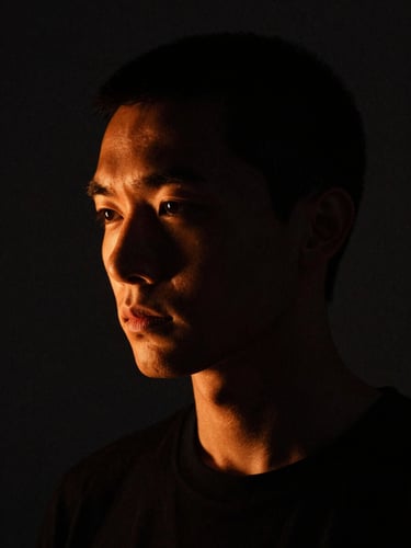 High-contrast portrait of the artist in a dark studio, single-source side lighting with warm orange rim light, dramatic chiaroscuro, deep charcoal tones, 35mm film grain