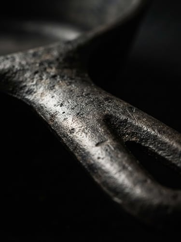 Extreme close-up macro shot of a heavy unlacquered iron skillet handle, dramatic side-lighting, deep shadows, dark moody background, high-contrast tactile metal textures