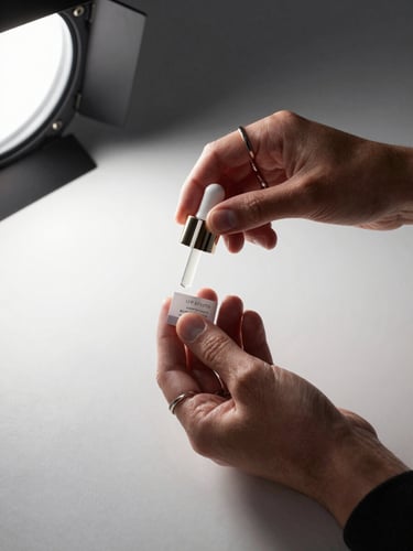 Close-up editorial frame: a creator's hands holding a skincare product over a minimal white surface, studio strobe light from the upper left casting a sharp shadow, product label in sharp focus, creator's sleeve and rings visible — a controlled brand-collaboration evidence shot, no face visible
