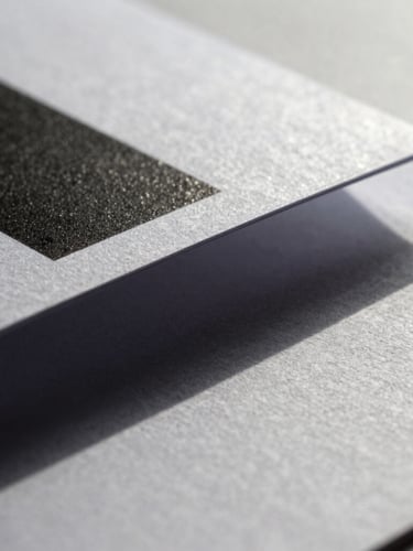 Macro crop of high-contrast printed brand collateral showing textured paper grain and a sharp geometric shadow cast under cool indirect daylight.