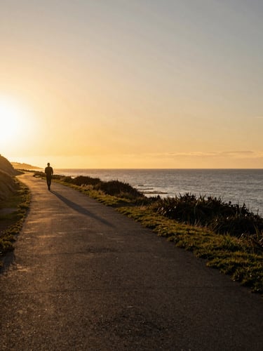 Wide environmental shot looking left along a sun-drenched coastal path, a lone figure silhouetted against the golden-hour horizon, open sea stretching far right, warm amber light raking across the ground
