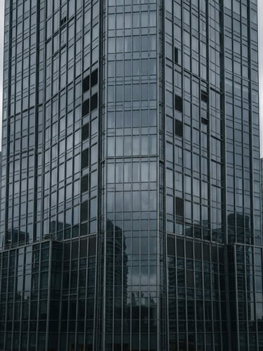 Wide architectural shot of a modern high-rise building facade, glazed curtain walls reflecting an overcast sky, cool desaturated steel and charcoal tones, sharp focus, 35mm
