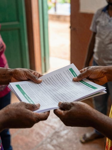 Close-up of hands distributing health supplies and printed wellness guides at a community gathering, natural daylight from a nearby open door, terracotta and green tones in the environment, focus on the exchange between two people