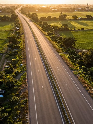 Wide-angle aerial drone photography of a newly paved multi-lane highway cutting through lush green Uttar Pradesh fields at golden hour, warm sunlight framing clean asphalt boundaries, high-contrast shadows