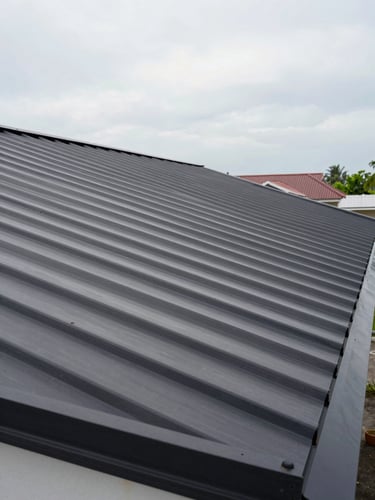 Wide environmental panoramic shot of a completed residential metal roof in Indonesia, natural overcast daylight, camera positioned low-right showing full roof span and ridge line, deep eaves visible on left, open sky above, no people