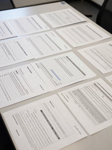 Wide overhead view of a policy audit workspace — structured documentation forms, classification grids, and annotated behavioral data laid flat on a large institutional table under even fluorescent light, papers precisely arranged, no people visible