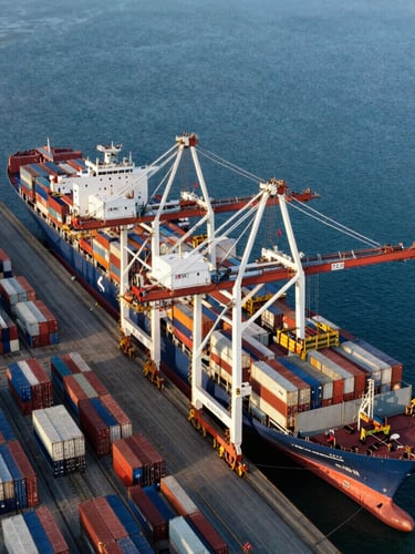 Overhead wide shot of a massive container terminal at golden hour, gantry cranes loading a mega container ship, deep blue water, clean geometric rows of colored containers.