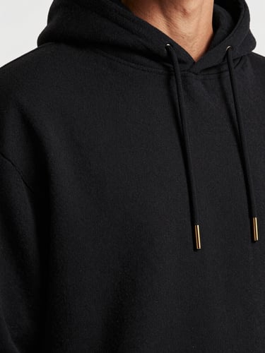Close-up macro shot of a heavy-drape black cotton hoodie on a model, soft natural shadows, highlighting the thick textile grain and precise stitching, desaturated tones, 35mm lens.