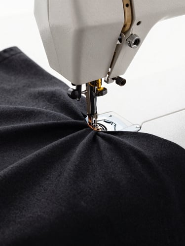 Macro close-up of heavy-drape black cotton fabric being stitched on an industrial sewing machine, bright diffused daylight highlighting the textile grain, premium editorial fashion photography style, 35mm lens