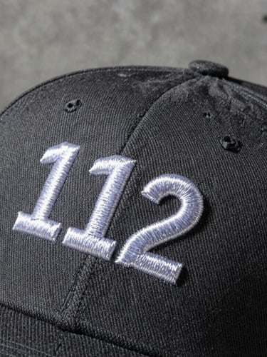 Macro tactile product photography of a structured charcoal Richardson 112 cap, high-contrast side-lighting highlighting the thick, high-density white embroidery stitches on the front crown, concrete workshop background.