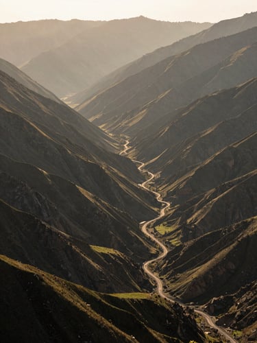 Environmental wide shot of a Himalayan valley at 4 PM, late afternoon light cutting across layered ridges, a winding unpaved track visible in the mid-ground, no people, muted green and ochre terrain, soft haze on distant peaks