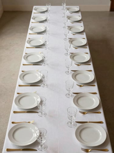 Overhead editorial shot of a long banquet table fully set for a formal event — white linens, precisely aligned charger plates, crystal glassware catching directional studio light, no guests, no faces, gold flatware in sharp focus against a cream surface, ultra-wide composition with generous negative space at top