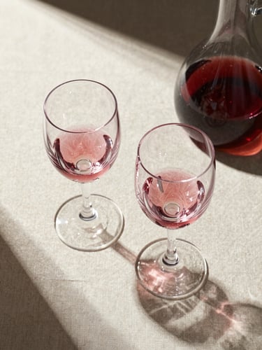 Overhead flat-lay of two crystal wine glasses and a decanter on a cream linen surface, wine catching directional studio light, sharp focus on the glass rim and liquid color, no people, minimal composition