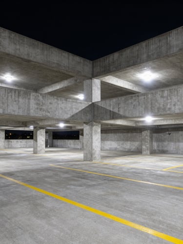 Wide-angle architectural shot of a multi-level concrete parking structure at night, stark high-contrast sodium lighting casting dramatic shadows, pristine clean concrete surface with bright yellow striping, no vehicles.