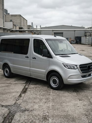 Three-quarter view of a clean modern silver passenger van parked in a desaturated industrial concrete yard, overcast daylight, crisp automotive photography, 35mm lens