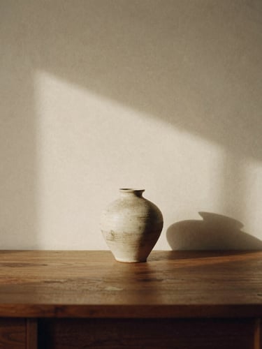 Stark minimalist workspace with warm light casting clean shadows on a textured cream wall, a singular ceramic vase on a dark oak desk, 35mm film grain, editorial composition.