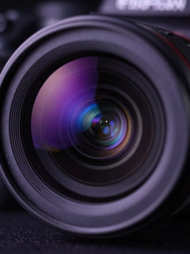 Extreme close-up of a professional camera lens in a dark studio setting, reflecting deep purple and vibrant blue key lights, cinematic shallow depth of field, subtle dust particles catching the light