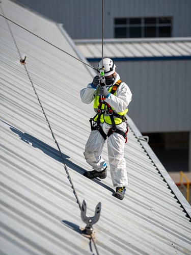 Close-up environmental shot of a technician in full PPE — harness, helmet, high-visibility vest — anchored to a lifeline cable on a steeply pitched industrial metal roof under harsh midday sun, structural corrugations casting sharp shadows, safety anchor point visible in foreground