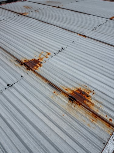 Close-up drone aerial of industrial corrugated metal roof showing rust staining and water infiltration damage annotated with visible markers, harsh overcast daylight creating strong texture on the metal surface, no workers visible, structural grid lines receding into the frame