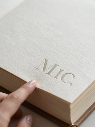 A close-up macro shot in soft, diffused morning light focusing on a hand gently turning the thick, textured paper page of a linen-bound memory album. Elegant, minimalist serif typography is embossed on the cover, showing rich paper grain and clean tactile edges.