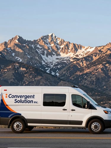 Wide panoramic view of the Colorado Front Range mountains at sunrise, with a branded Convergent Solution Inc. service vehicle parked on a highway shoulder in the foreground, facing right, crisp daylight.
