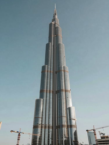 Wide environmental shot of an under-construction luxury tower in Dubai, steel and glass structure rising against a clear midday sky, construction cranes flanking the building, architectural scale emphasized