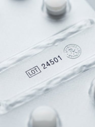 Macro shot of sealed pharmaceutical packaging with a clear lot number and regulatory stamp, sterile white and cool gray tones, bright studio lighting, sharp focus.