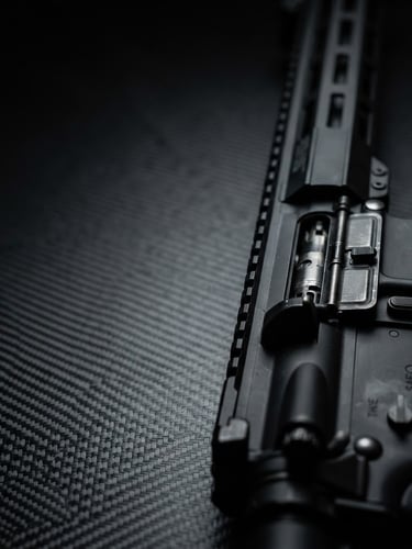 High-contrast macro shot of a modern black tactical rifle on a dark carbon fiber surface, sharp side-lighting, metallic highlights, 50mm lens, wide frame, right third.