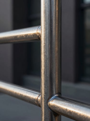 Extreme close-up of a stainless steel railing joint, shot from a low angle against a dark architectural wall — late-afternoon raking light catches the brushed metal grain and the precision weld seam, revealing surface texture and material quality; tight macro framing with shallow depth of field, no people