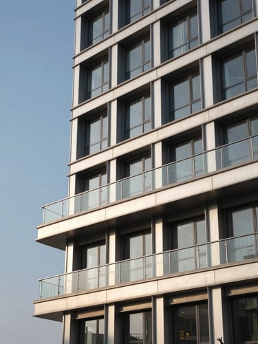 Wide environmental shot of a completed commercial building facade showing flush-fitted aluminium window frames and a continuous stainless steel glass railing on the upper balcony, late-afternoon directional light casting long shadows that reveal the precision of the frame reveals, clean architectural lines, no people, building occupies right two-thirds of frame with open sky to the left