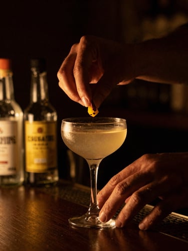 Close-up of a mixologist's hands delicately placing a garnish on a cocktail, house-made bitters bottles in background, warm golden light, dark moody tones, shallow depth of field.